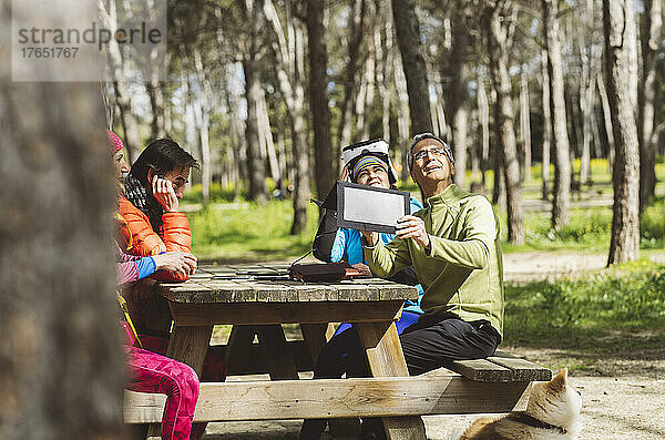 Reifer Mann passt Solarpanel unter Sonnenlicht an und sitzt mit Freunden am Picknicktisch im Wald