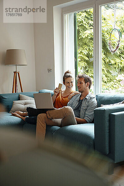 Glückliche Frau mit Kreditkarte sitzt neben ihrem Freund und benutzt Laptop  der zu Hause auf dem Sofa sitzt