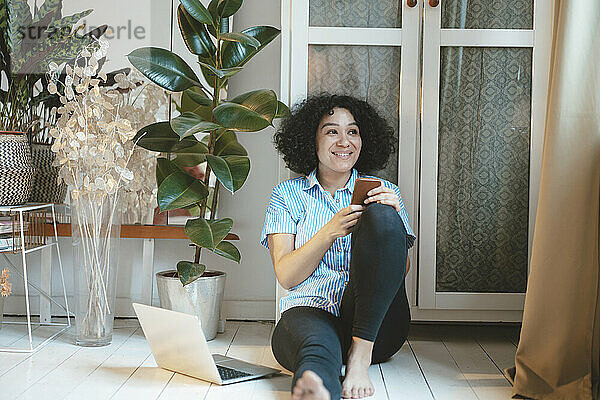 Lächelnde Frau mit Mobiltelefon sitzt zu Hause am Laptop