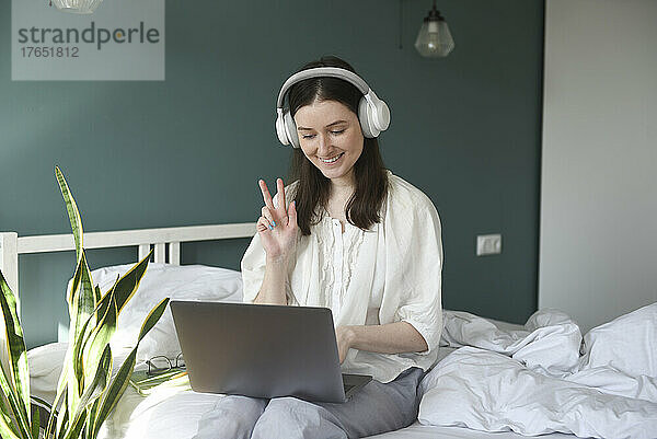 Glückliche Frau mit kabellosen Kopfhörern gestikuliert Friedenszeichen und sitzt mit Laptop auf dem Bett