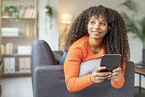Lächelnde Frau mit Smartphone auf Sofa im Wohnzimmer zu Hause