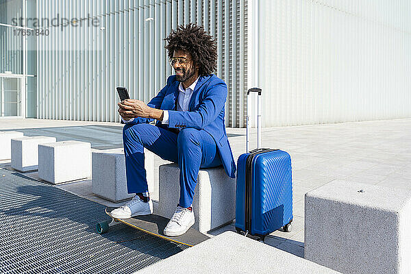 Geschäftsmann mit Koffer und Skateboard mit Mobiltelefon sitzt auf Betonblock