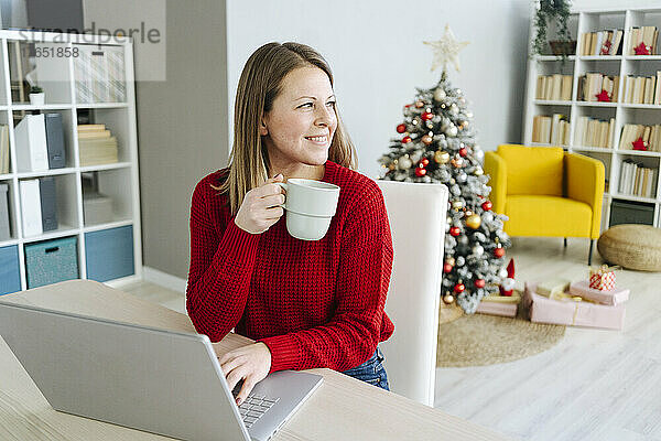 Lächelnde Frau mit Kaffeetasse und Laptop sitzt am Tisch im Wohnzimmer