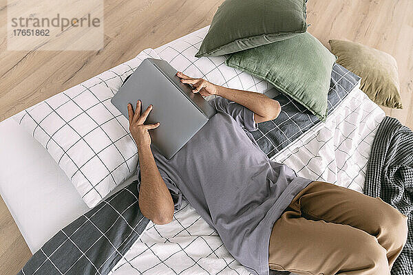 Young man covering face with laptop lying on bedding at home