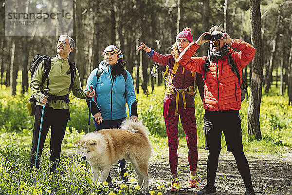 Mann schaut durch ein Fernglas und steht neben Freunden und Hund im Wald