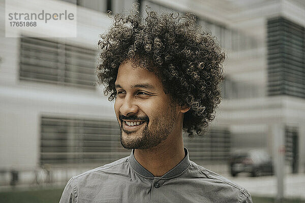 Lächelnder junger Mann mit lockigem Haar vor einem modernen Gebäude