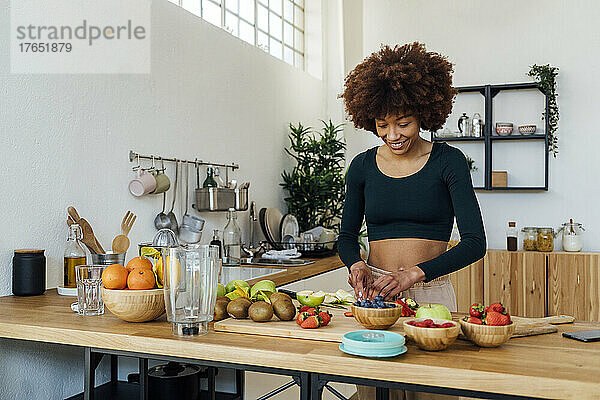 Lächelnde junge Frau schneidet Früchte und steht am Tisch in der Küche
