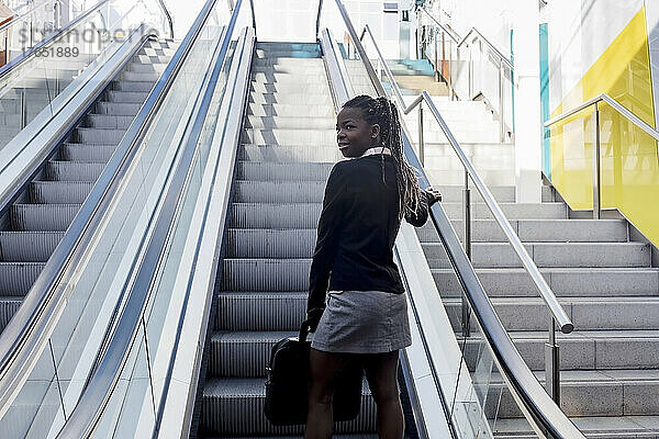 Lächelnde junge Frau mit Laptoptasche steht auf Rolltreppe