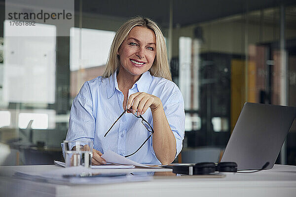 Lächelnde Geschäftsfrau mit Papier und Brille sitzt am Laptop am Schreibtisch im Büro