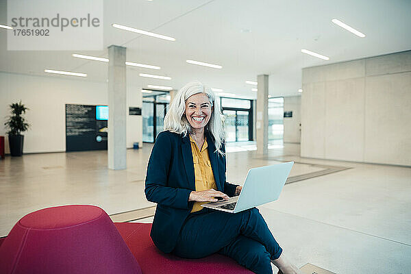 Cheerful businesswoman with laptop sitting on seat in office
