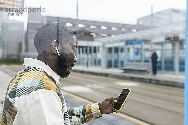 Mann hört Musik über kabellose In-Ear-Kopfhörer mit Mobiltelefon an der Straßenbahnhaltestelle