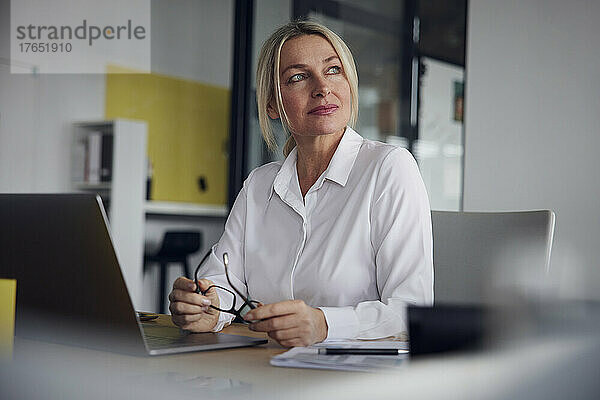 Geschäftsfrau hält Brille und Laptop am Schreibtisch im Büro
