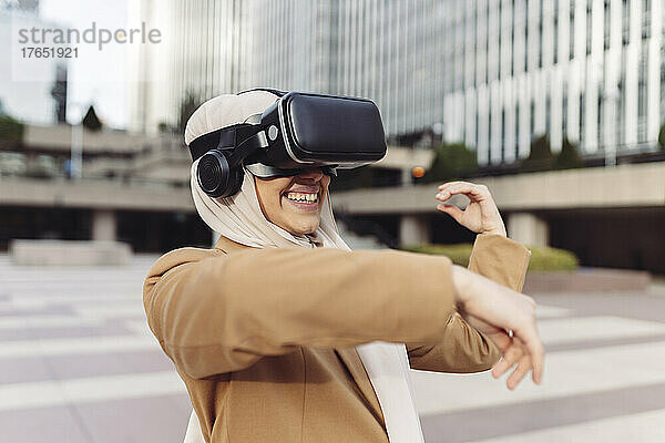Glückliche junge Geschäftsfrau mit Virtual-Reality-Simulator gestikuliert an einem sonnigen Tag im Büropark