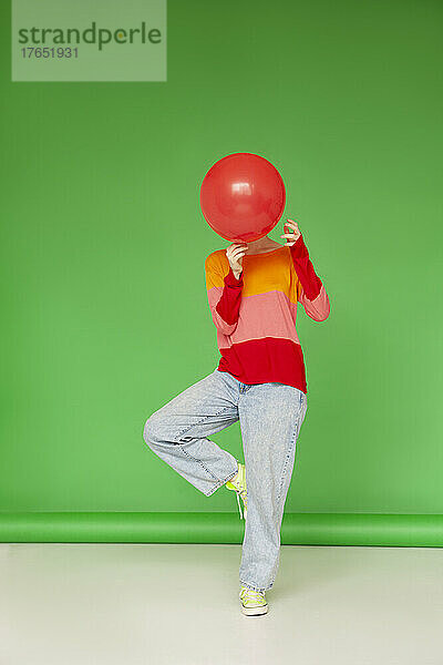 Frau bedeckt ihr Gesicht mit einem roten Ballon  der auf einem Bein vor grünem Hintergrund steht