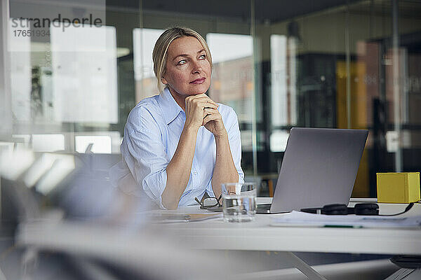 Nachdenkliche Geschäftsfrau mit der Hand am Kinn sitzt am Laptop am Schreibtisch im Büro