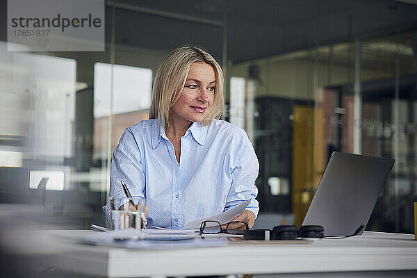 Geschäftsfrau blickt auf Laptop und schreibt am Schreibtisch im Büro in Papier