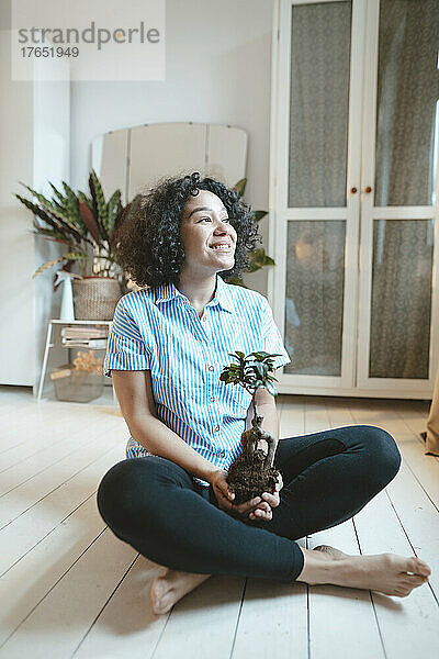 Lächelnde Frau sitzt zu Hause mit Zimmerpflanze auf dem Boden