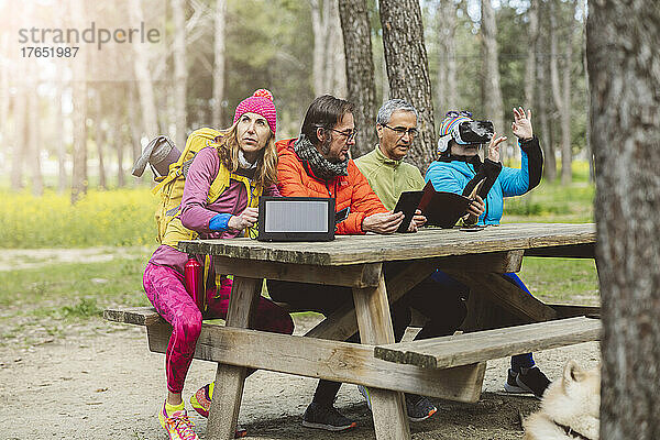 Reifer Mann mit Solarpanel sitzt bei Freunden am Picknicktisch im Wald