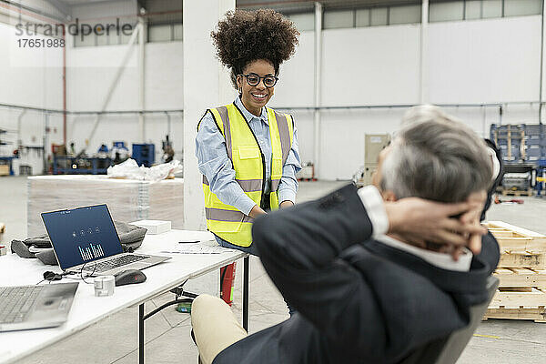 Lächelnder Ingenieur blickt Geschäftsmann an  der mit den Händen hinter dem Kopf in der Fabrik sitzt