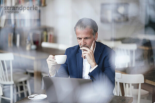 Geschäftsmann mit Hand am Kinn und Kaffeetasse mit Laptop im Café