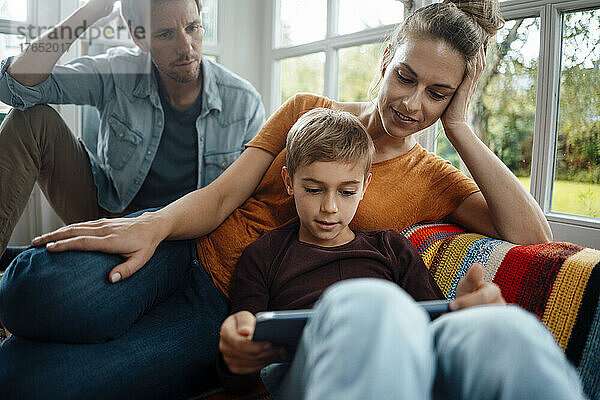 Mann und Frau schauen Sohn mit Tablet-PC an  der zu Hause auf dem Sofa sitzt