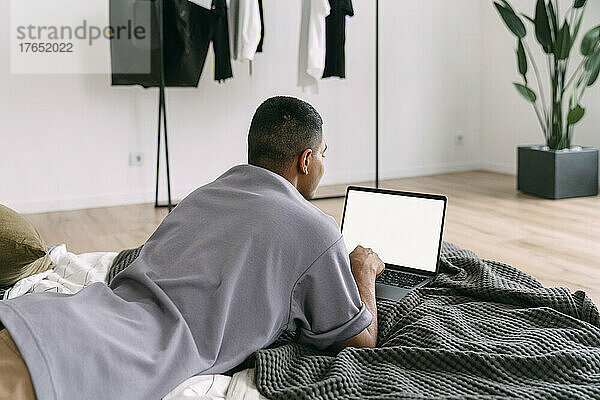 Junger Mann benutzt Laptop und liegt zu Hause auf dem Bett