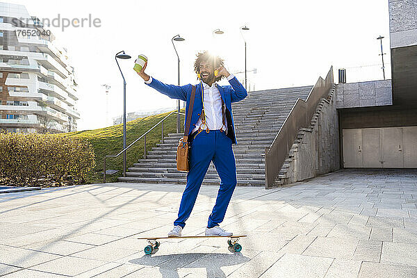 Glücklicher Geschäftsmann hält Einwegbecher in der Hand  hört Musik und fährt Skateboard auf dem Fußweg