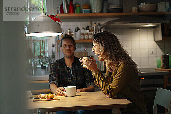 Fröhliche Frau hält Kaffeetasse in der Hand und sitzt mit ihrem Freund am Esstisch in der Küche