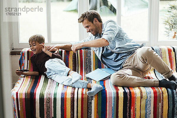 Glücklicher Vater blickt auf verspielten Sohn  der zu Hause auf dem Sofa sitzt und sein Smartphone benutzt
