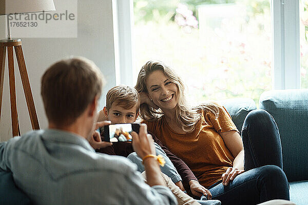 Glückliche blonde Frau und Sohn  fotografiert von Mann per Smartphone im Wohnzimmer