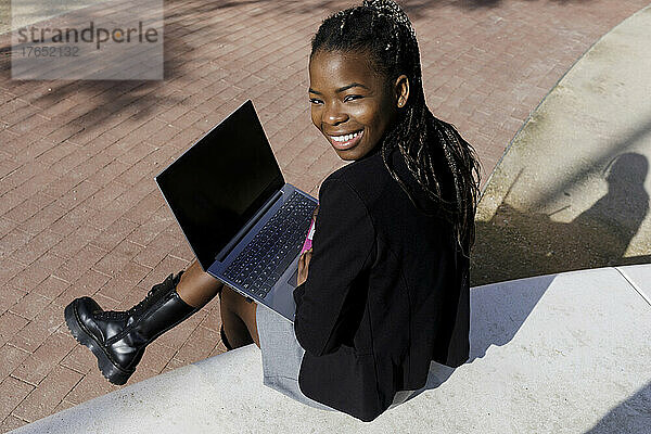 Glückliche junge Frau mit Laptop sitzt auf Betonsitz