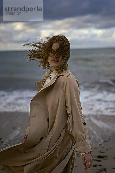 Frau mit zerzausten Haaren trägt Trenchcoat am Strand