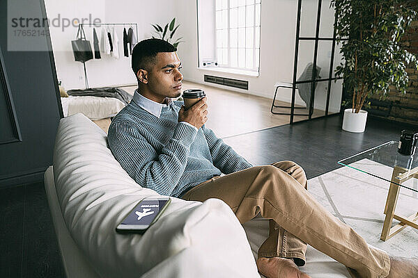 Mann mit Einwegkaffeetasse sitzt neben Smartphone und Flugmodussymbol auf dem heimischen Sofa