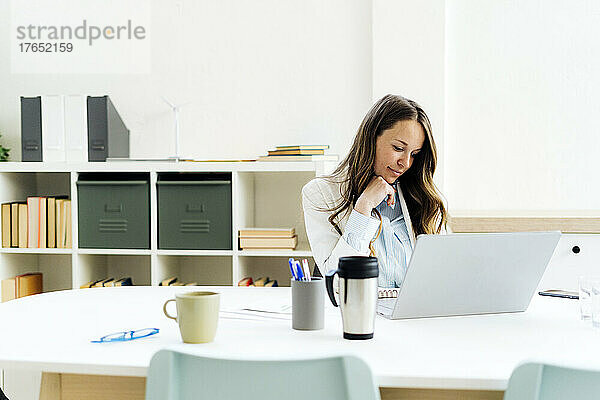 Geschäftsfrau mit Laptop sitzt mit der Hand am Kinn am Schreibtisch