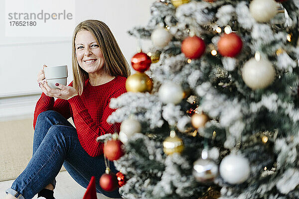 Glückliche Frau mit Kaffeetasse sitzt zu Hause am Weihnachtsbaum