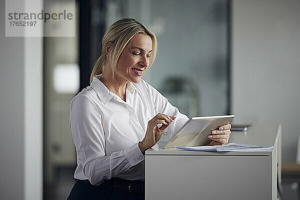 Lächelnde Geschäftsfrau mit Tablet-PC  die neben dem Schrank im Büro steht