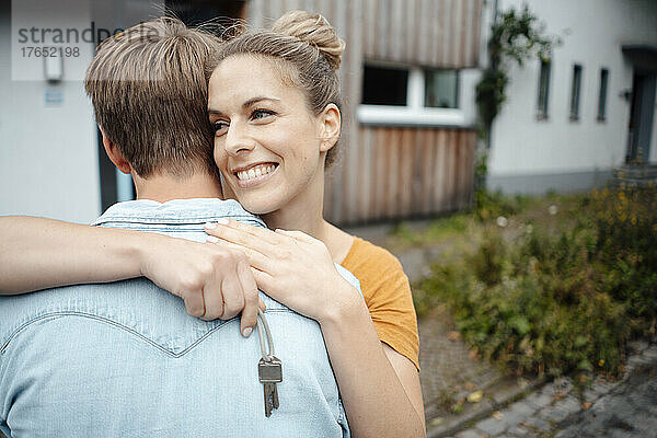 Glückliche blonde Frau hält den Hausschlüssel in der Hand und umarmt ihren Freund im Hinterhof