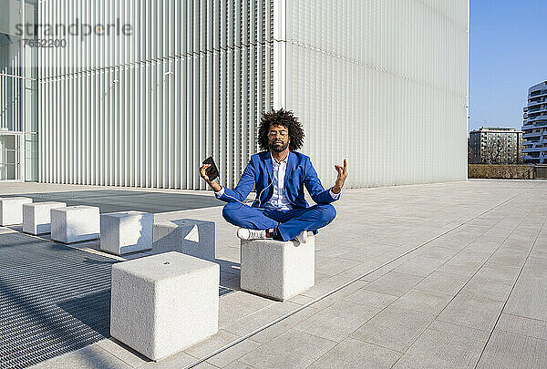 Geschäftsmann mit Mobiltelefon hört Musik und meditiert auf Betonblock