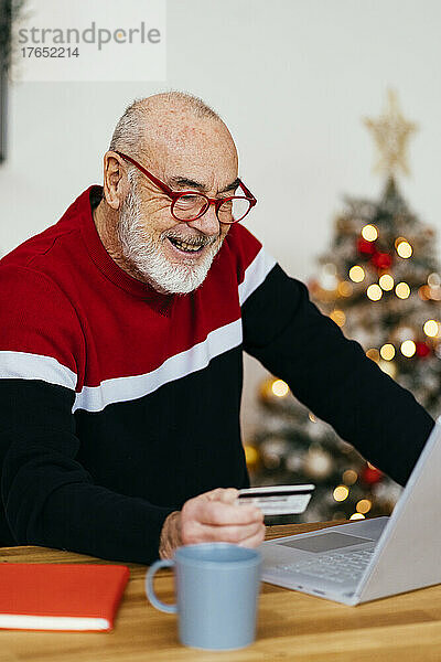 Glücklicher älterer Mann mit Kreditkarte  der zu Hause über den Laptop online einkauft