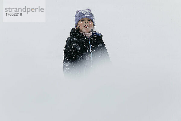 Mädchen mit Strickmütze streckt die Zunge heraus und steht im Schnee