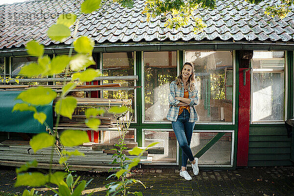 Glückliche Frau steht mit verschränkten Armen vor einem Glasfenster im Hinterhof