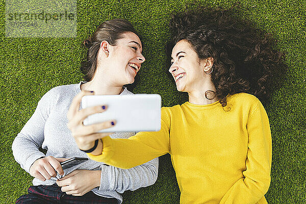 Cheerful friends taking selfie through smart phone lying down on grass at park