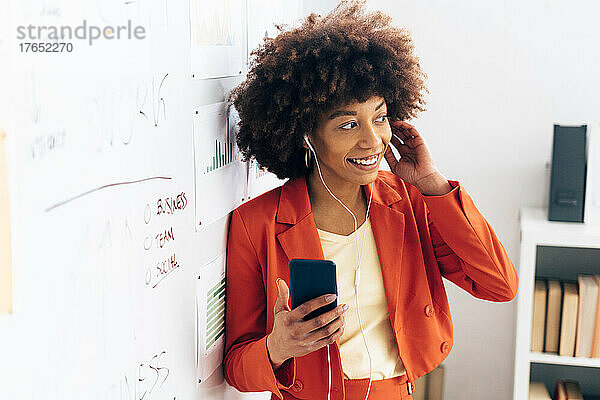 Lächelnde Geschäftsfrau hält Smartphone in der Hand und hört im Büro Musik über In-Ear-Kopfhörer