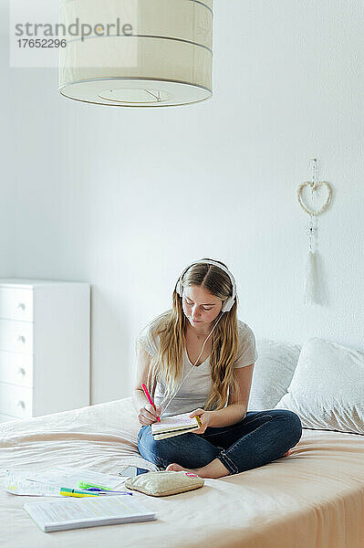Junge Frau mit Kopfhörern macht Hausaufgaben und sitzt zu Hause im Bett
