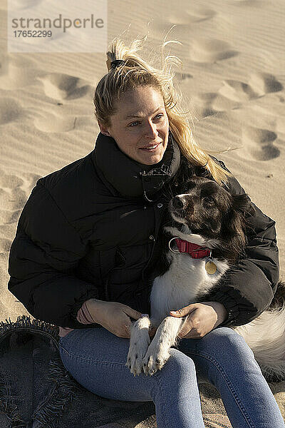 Lächelnde junge blonde Frau sitzt mit Hund im Sand