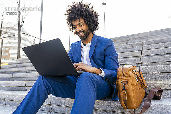Lächelnder Geschäftsmann mit Laptop  der neben der Umhängetasche auf der Treppe sitzt