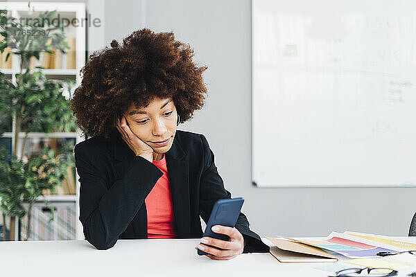 Gelangweilte Geschäftsfrau sitzt am Schreibtisch mit der Hand am Kinn und benutzt das Mobiltelefon im Büro