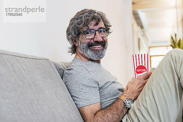 Glücklicher älterer Mann mit Popcorn  der zu Hause auf dem Sofa sitzt