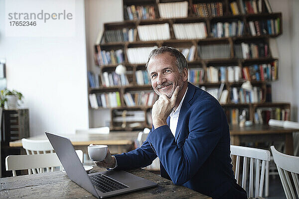 Lächelnder Geschäftsmann mit Kaffeetasse und Laptop sitzt am Tisch im Café