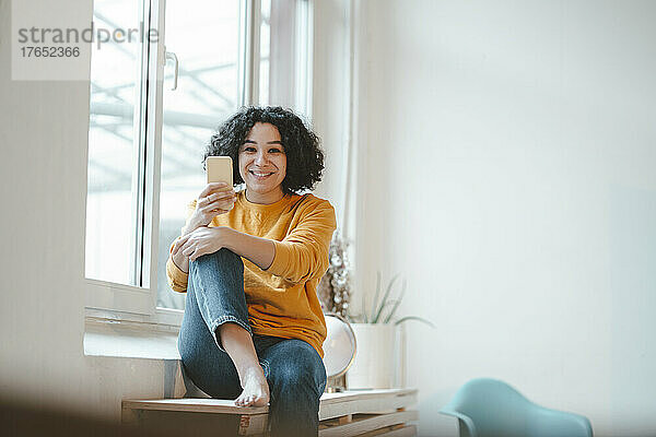 Lächelnde Frau mit Mobiltelefon sitzt zu Hause am Tisch am Fenster